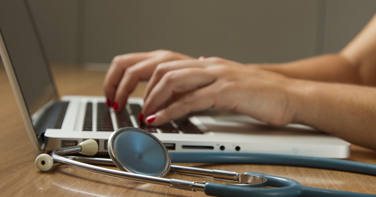 Mãos digitando em um laptop ao lado de um estetoscópio, representando tecnologia na medicina